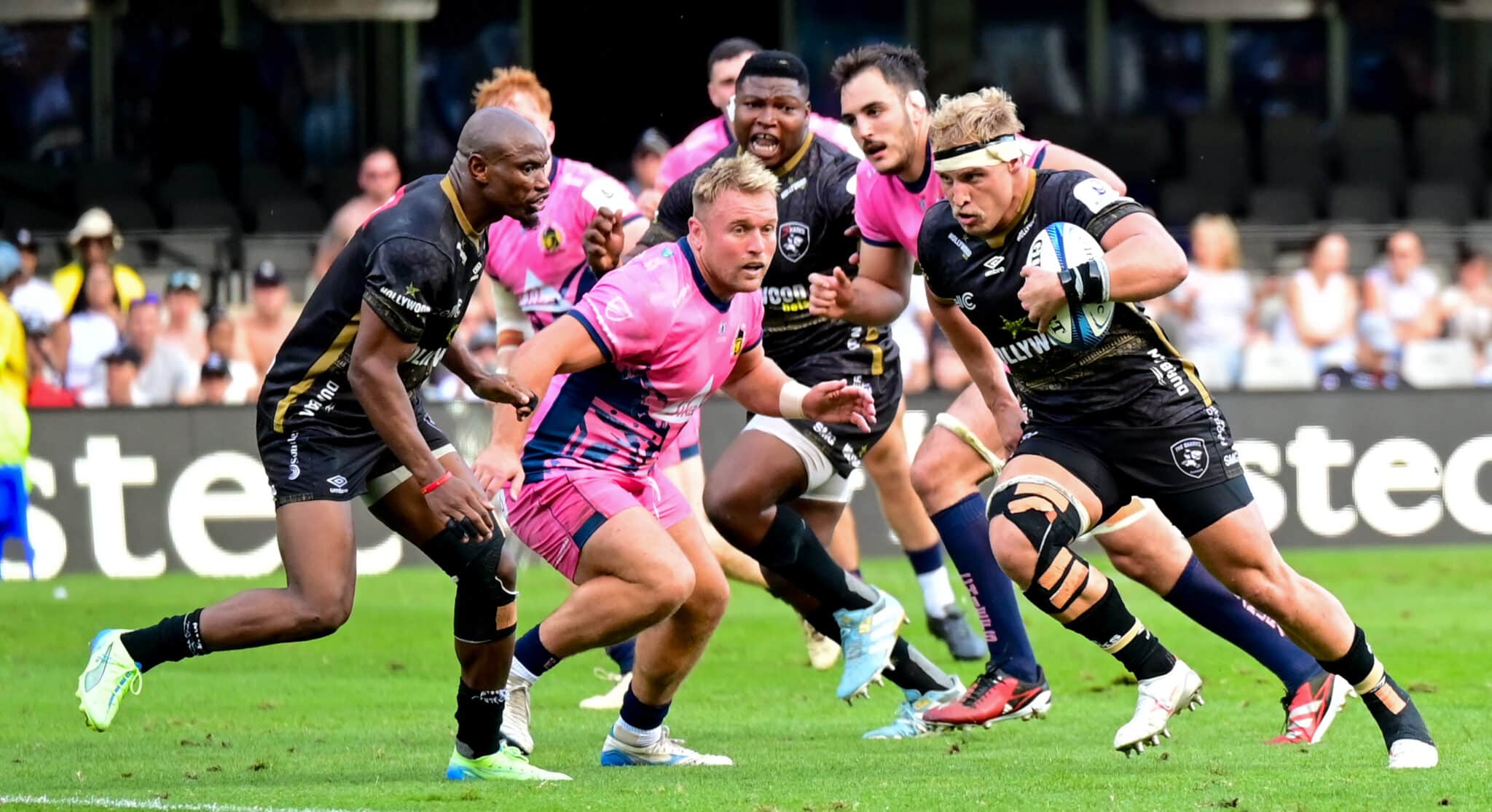 In the image, a dynamic moment from a rugby match is captured, showcasing a player in a vibrant pink kit, driving forward with the ball against the opposition. Surrounding him are players from the opposing team, dressed in black kits, who are intensely focused on halting his advance. The intensity and focus of the players are palpable, highlighting the physicality and fast-paced action typical of a high-stakes rugby game. This scene perfectly illustrates the competitive spirit and athleticism inherent in the sport.