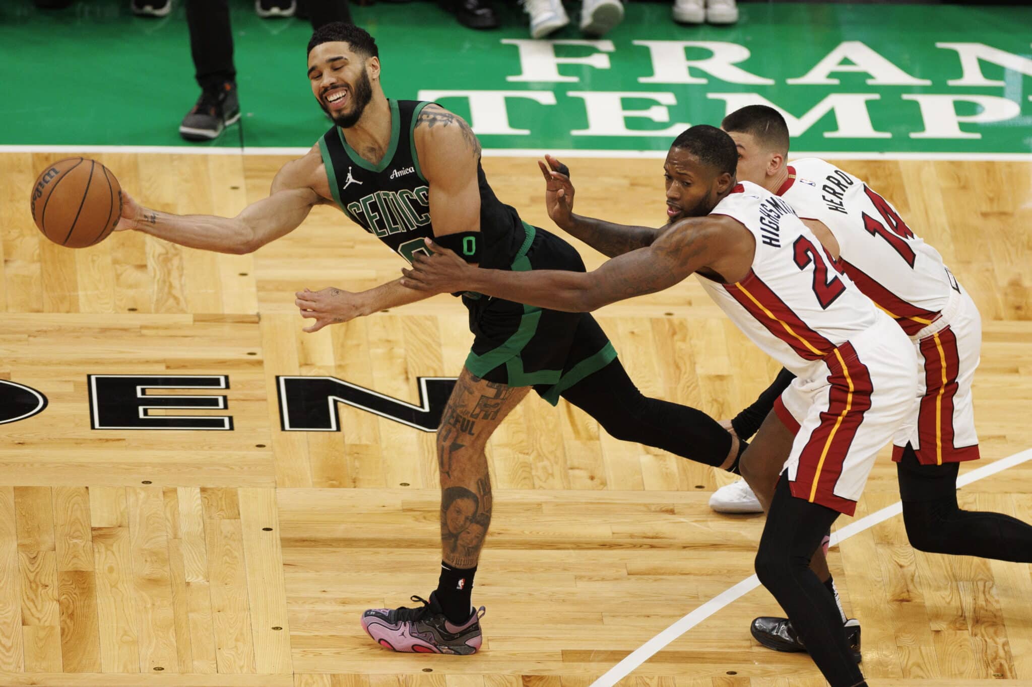 This action-packed image captures a dynamic moment during a basketball game, featuring a player from the Boston Celtics driving towards the basket with the ball, while defenders from the Miami Heat try to block his path. The Celtics player, wearing a green jersey with the number 0, exhibits skillful control and determination as he maneuvers around his opponents. The intense focus and physicality of the players are vividly displayed against the backdrop of the vibrant court, adding to the excitement of the match.