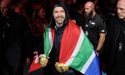 A South African MMA fighter draped in the national flag, wearing gold gloves, smiles as he makes his entrance at a UFC event, surrounded by his team and security.