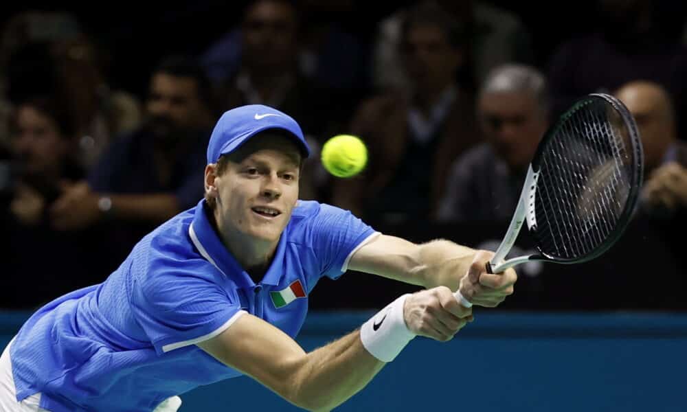 This image captures a tennis player in intense action during a match, dressed in a bright blue shirt with a national flag emblem, suggesting he's playing in a significant international event. He is focused on a low, forceful backhand stroke, showing excellent form and concentration. The blurred background emphasizes the speed and dynamism of the game, highlighting the player's athletic prowess and the high stakes of the competition.