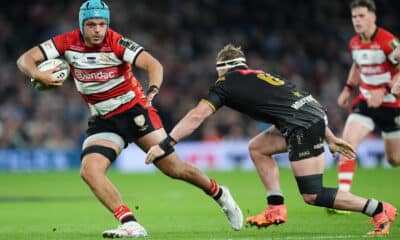 A rugby player in a red and white jersey, identified as a member of the Gloucester Rugby team, runs powerfully with the ball, fending off a tackle from an opposing Exeter Chiefs player in a black and gold uniform during a match.