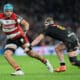 A rugby player in a red and white jersey, identified as a member of the Gloucester Rugby team, runs powerfully with the ball, fending off a tackle from an opposing Exeter Chiefs player in a black and gold uniform during a match.