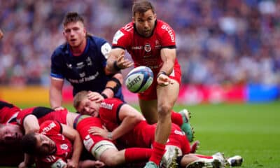 A rugby player in a red jersey from Toulouse deftly passes the ball during a dynamic play, as opponents from Leinster in navy blue attempt to tackle him, showcasing intense action on the rugby field.