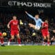 The image captures a dramatic moment from a football match, possibly between Manchester City and a team in red, judging by the uniforms. A player in Manchester City's light blue kit performs an acrobatic kick to play the ball, surrounded by opposing players in red. This action-packed scene occurs under the floodlights of Etihad Stadium, indicating an evening game filled with high energy and athleticism. Such images are emblematic of the intense competition in top-tier football matches, where every move can significantly impact the game's outcome.