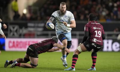 This image captures an intense moment from a rugby match. The player in the white jersey, showcasing determination, is running with the ball, while two opponents in black and pink-striped jerseys attempt to tackle him. The scene is a vivid display of the physicality and dynamic action typical of rugby, highlighting the players' focus and athleticism during the game.