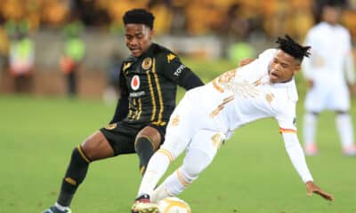 Two soccer players are competing fiercely for the ball during a match. One player, dressed in a black and gold uniform, is maneuvering to control the ball while the other, in a white kit with gold accents, is attempting a tackle or block.