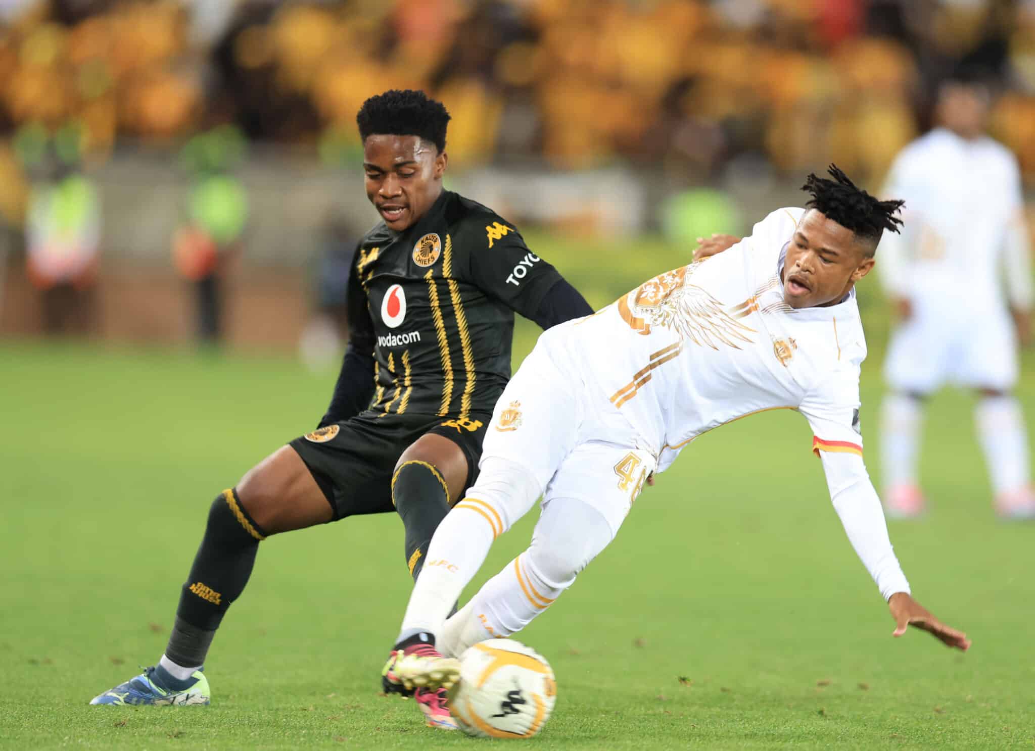 Two soccer players are competing fiercely for the ball during a match. One player, dressed in a black and gold uniform, is maneuvering to control the ball while the other, in a white kit with gold accents, is attempting a tackle or block.