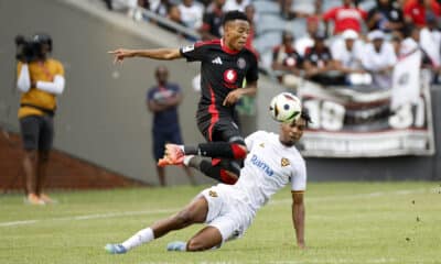 A dynamic action shot from a soccer match showing a player from Orlando Pirates in black and red gear jumping to head the ball, while a player from a team in white with a gold emblem attempts a tackle on the ground, with spectators and a cameraman in the background.