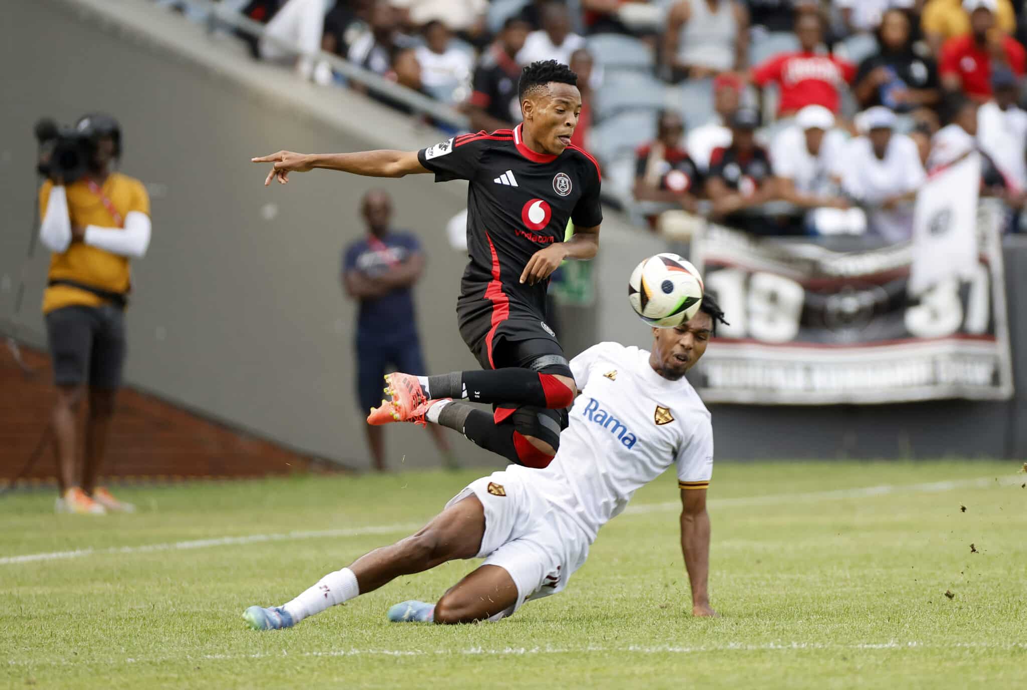 A dynamic action shot from a soccer match showing a player from Orlando Pirates in black and red gear jumping to head the ball, while a player from a team in white with a gold emblem attempts a tackle on the ground, with spectators and a cameraman in the background.