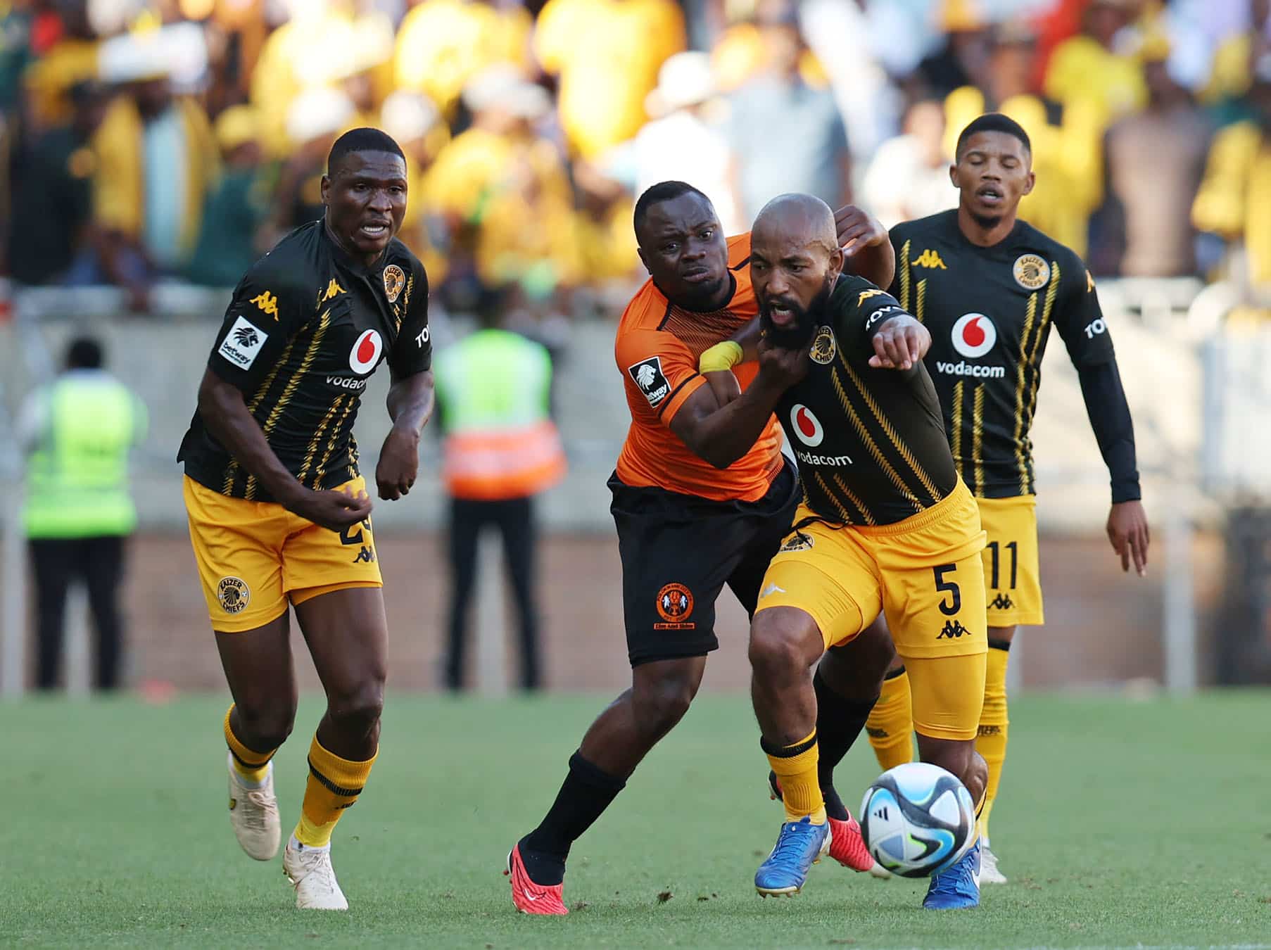 In the image, a moment from a soccer match between two teams, one in black and gold uniforms and the other in orange, captures a tense competition for the ball. The player in orange is tightly defending against a player from the opposing team, who is making a determined effort to maintain possession. This scene vividly illustrates the physical and strategic elements of soccer, showcasing the players' intense focus and athletic skill during a crucial match play.